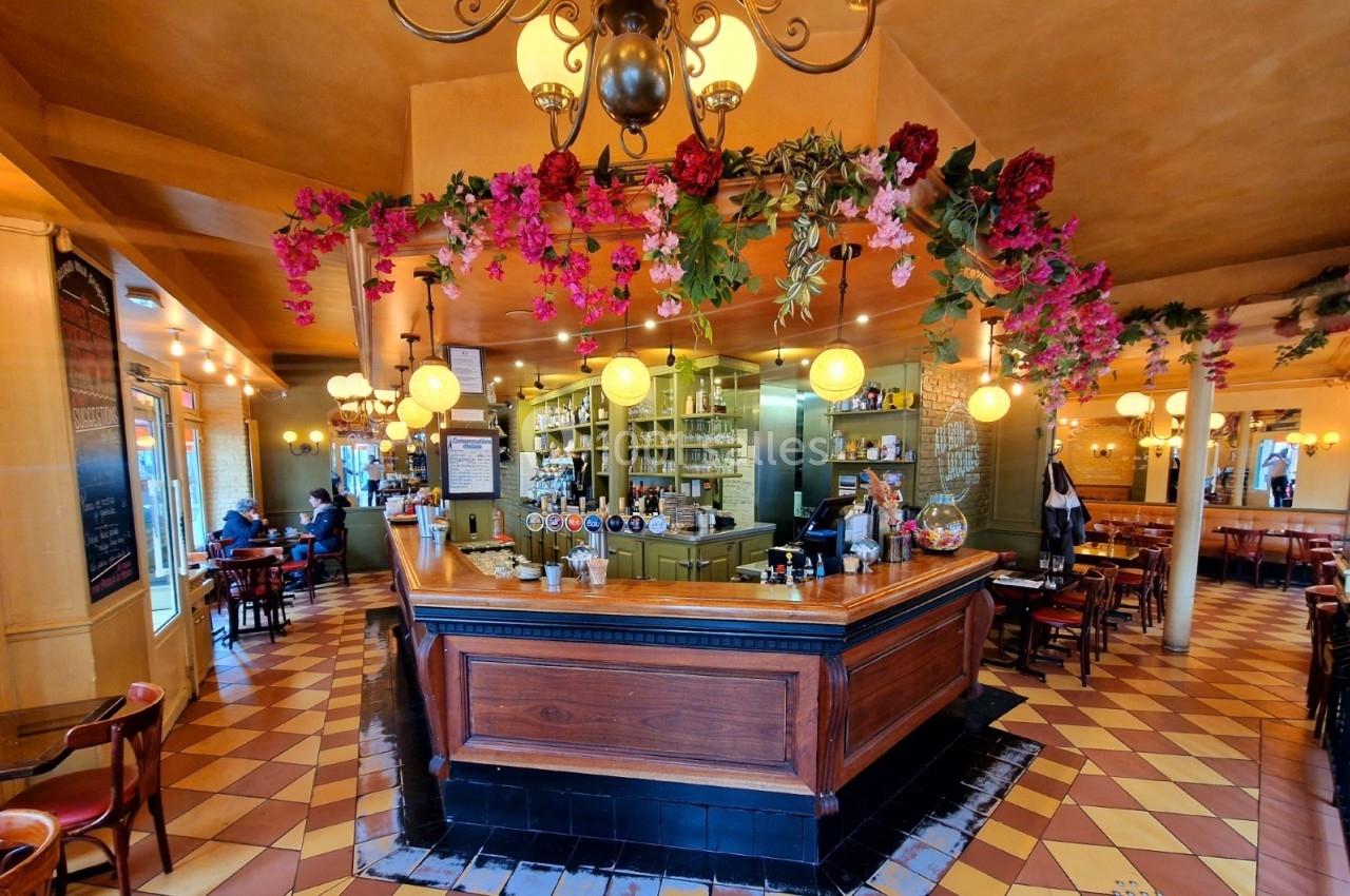 Intérieur d'un café avec un bar en bois central, décoré de fleurs suspendues et éclairé par des luminaires ronds.