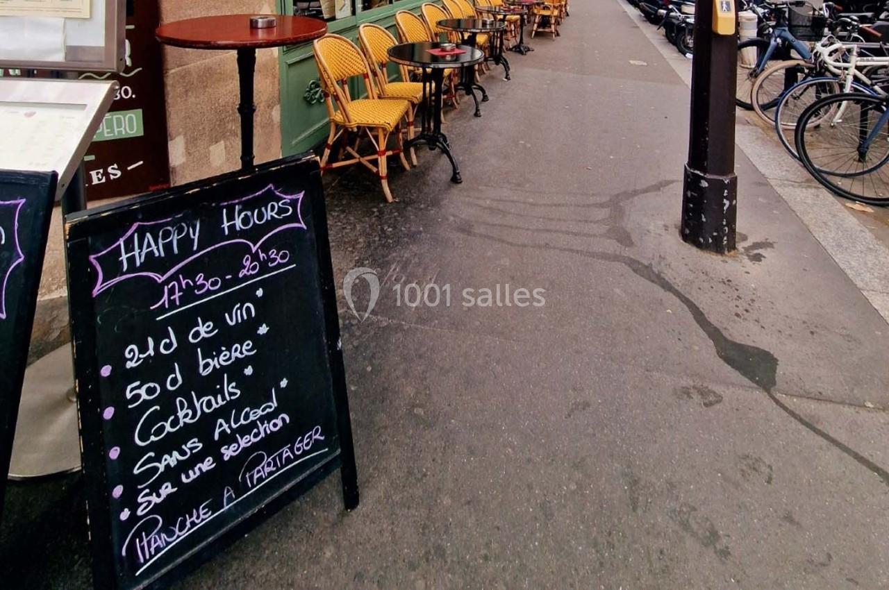 Ardoise indiquant les offres Happy Hours devant une terrasse de café avec chaises jaunes sur un trottoir urbain.