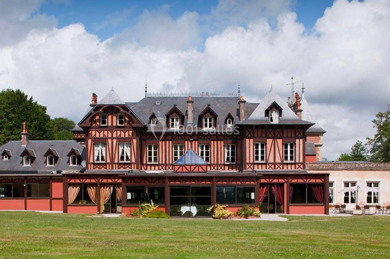 Façade d'un grand bâtiment à colombages rouges et noirs, entouré d'un jardin verdoyant sous un ciel nuageux.