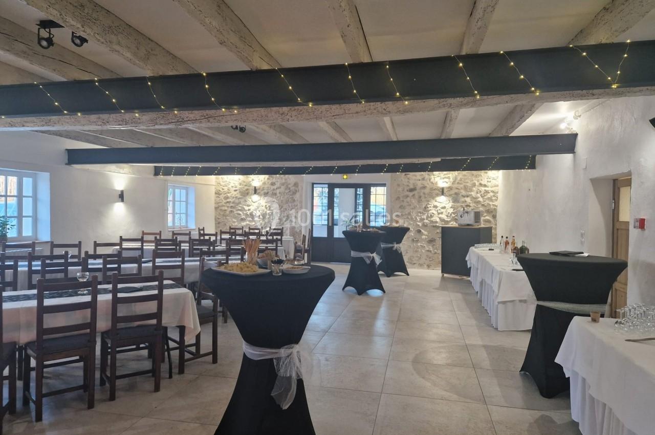 Salle de réception avec tables dressées, chaises en bois, poutres apparentes et guirlandes lumineuses au plafond.