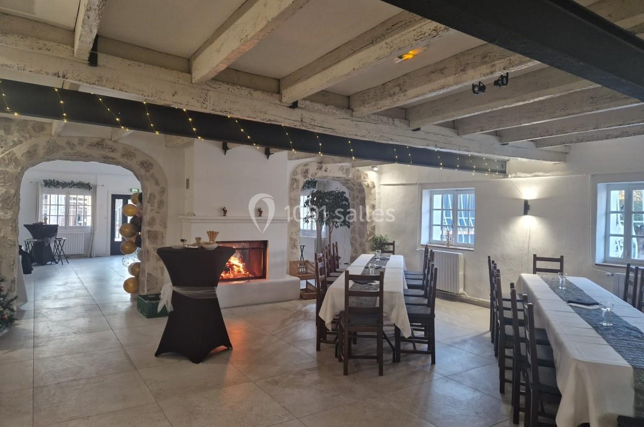 Salle lumineuse avec poutres apparentes, tables dressées, cheminée allumée et décorations discrètes.