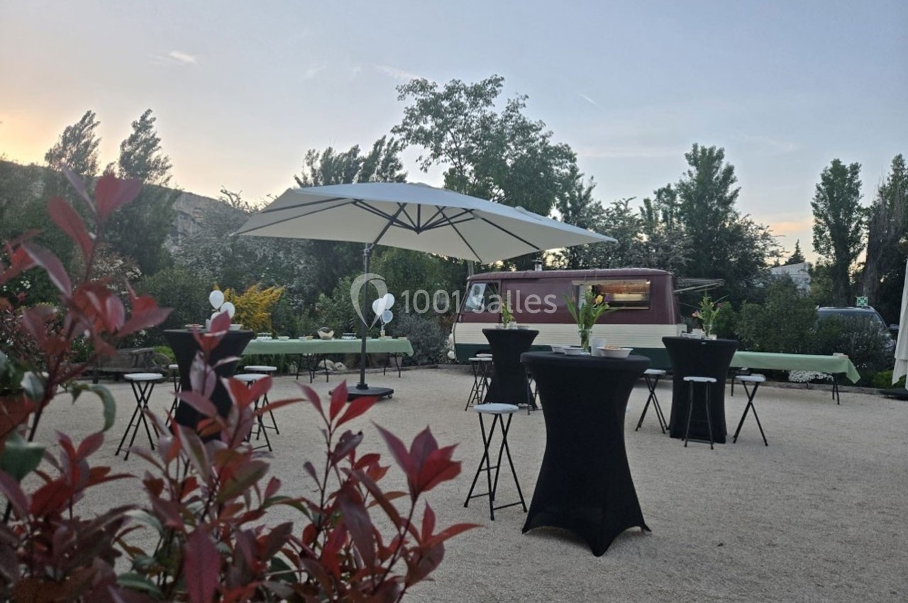 Espace extérieur aménagé avec tables hautes, parasols et un food truck, entouré de végétation au crépuscule.