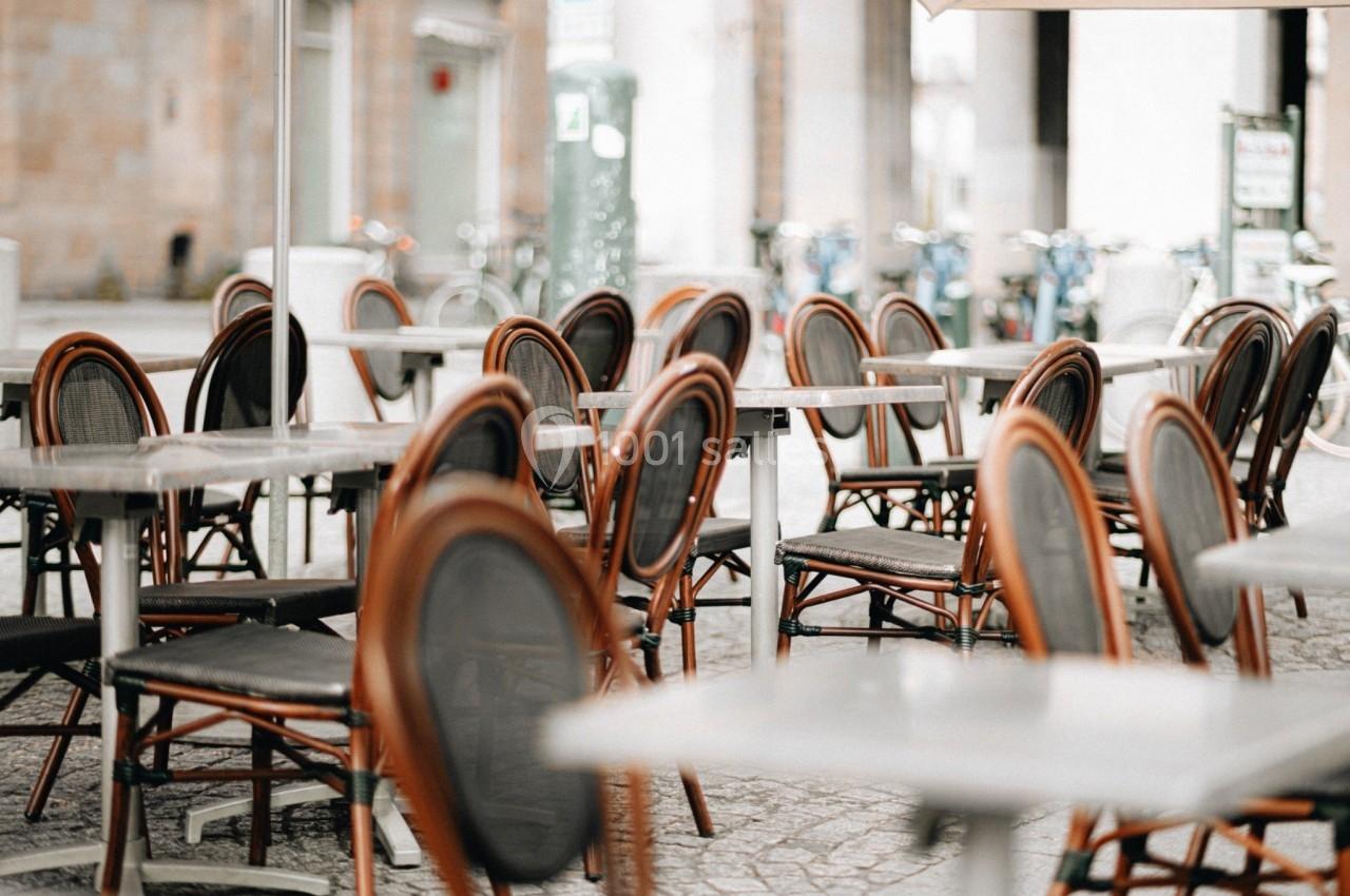 Chaises et tables vides disposées en terrasse extérieure sur une place pavée.