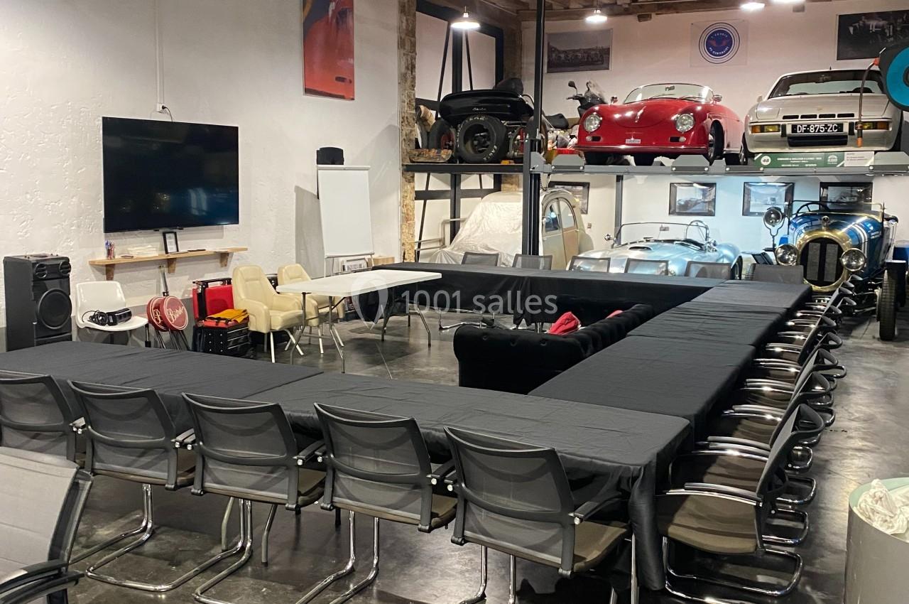 Salle de réunion aménagée dans un garage avec des voitures exposées en arrière-plan et des tables disposées en U.