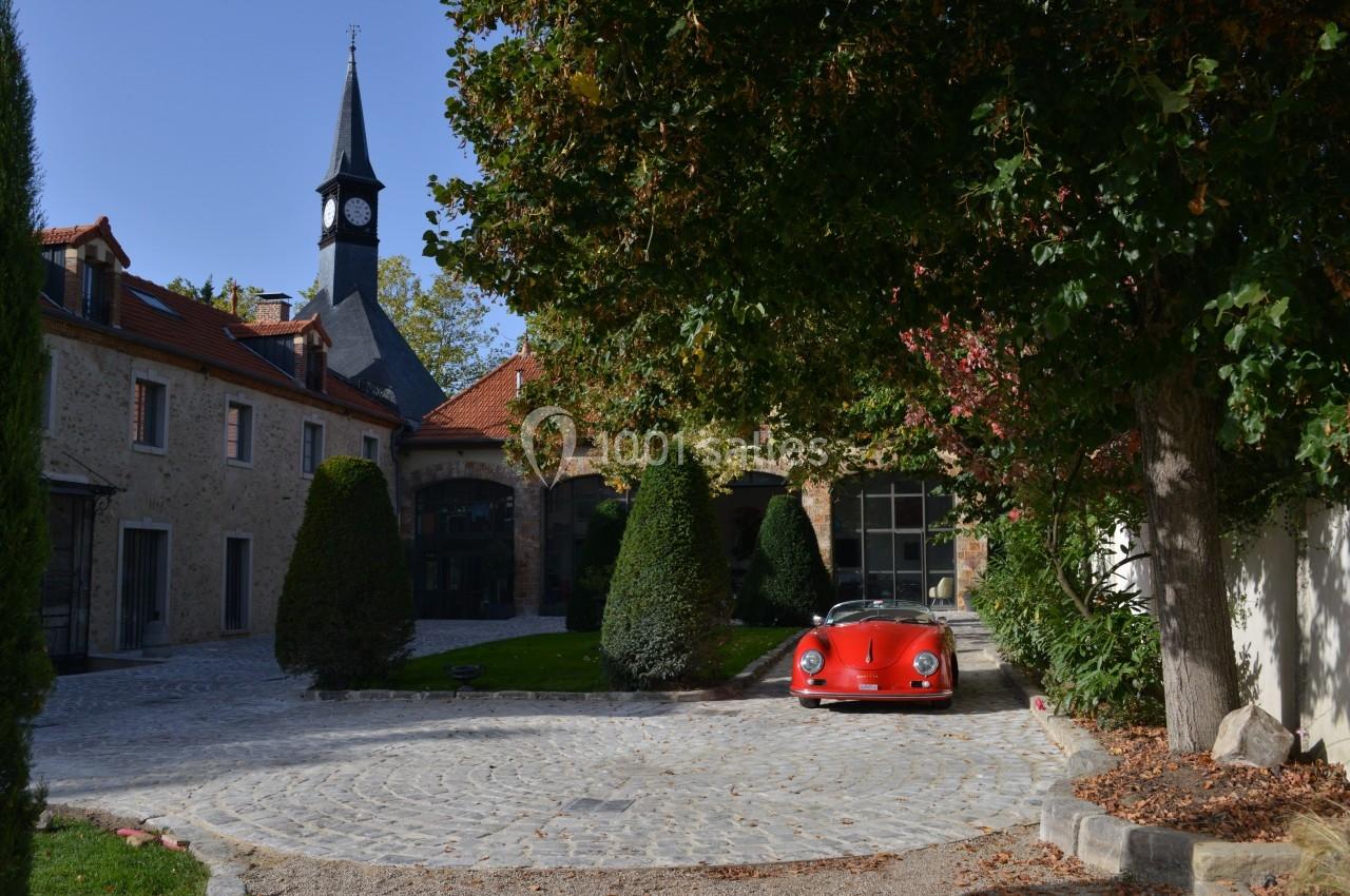 Voiture rouge garée sur une cour pavée entourée de bâtiments en pierre, avec un clocher visible à l'arrière-plan.
