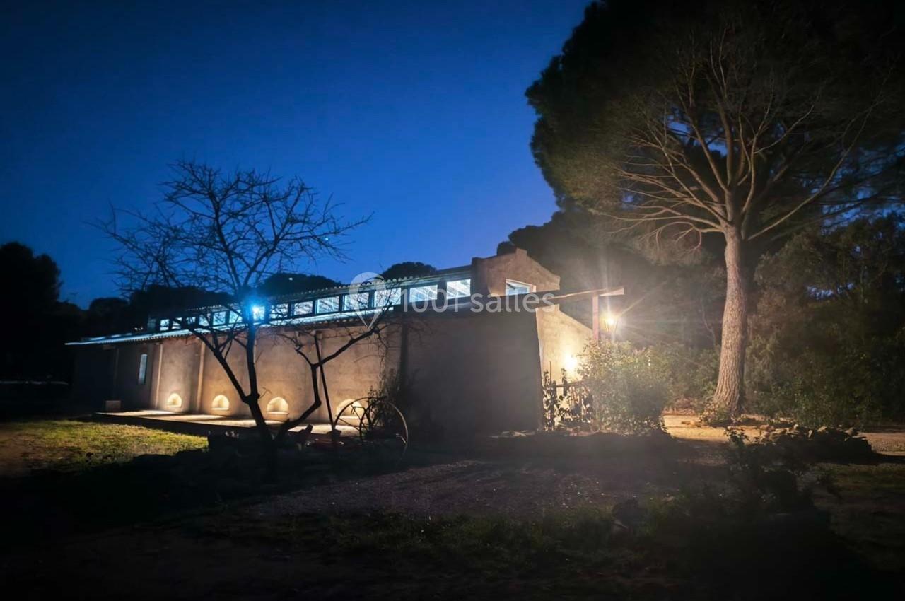 Maison moderne éclairée la nuit, entourée d'arbres et d'un jardin, avec un ciel sombre en arrière-plan.