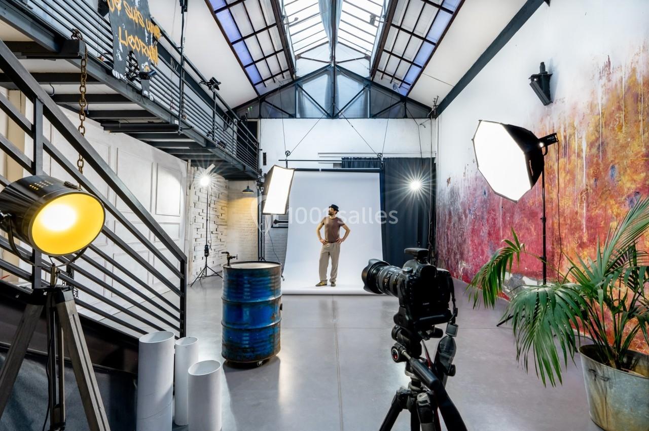 Studio photo lumineux avec un modèle debout devant un fond blanc, entouré d'équipements d'éclairage et d'une caméra.