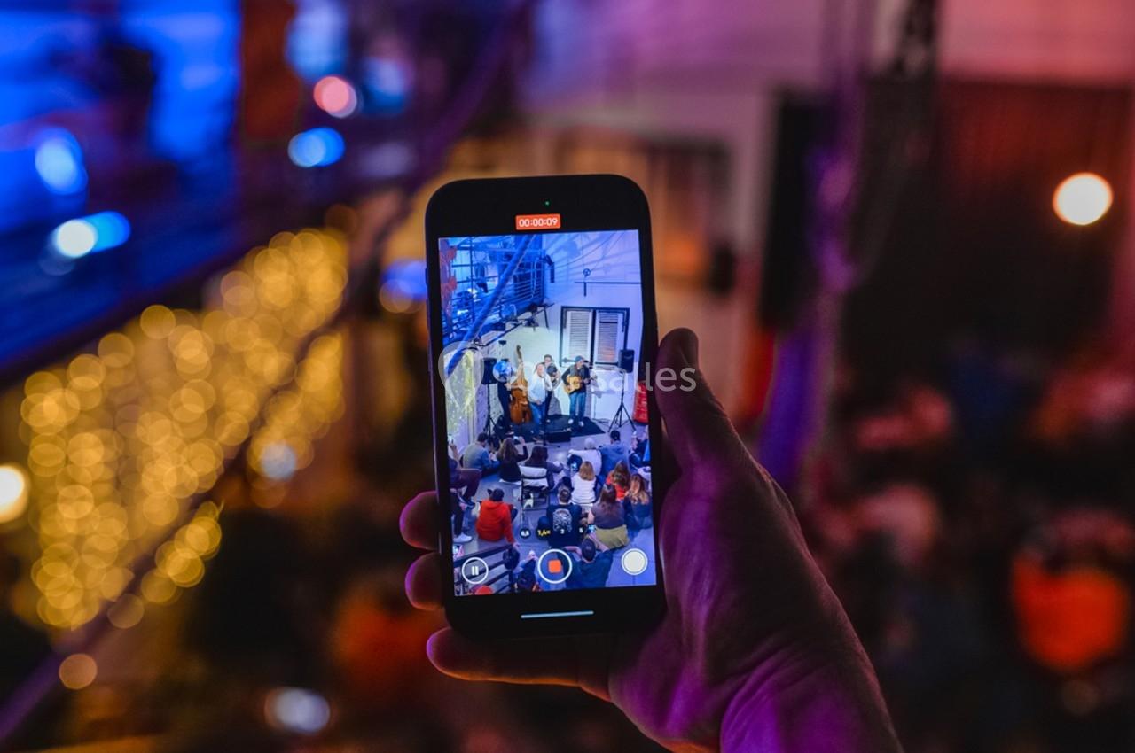 Une main tient un smartphone filmant un concert dans un lieu éclairé par des lumières colorées et tamisées.