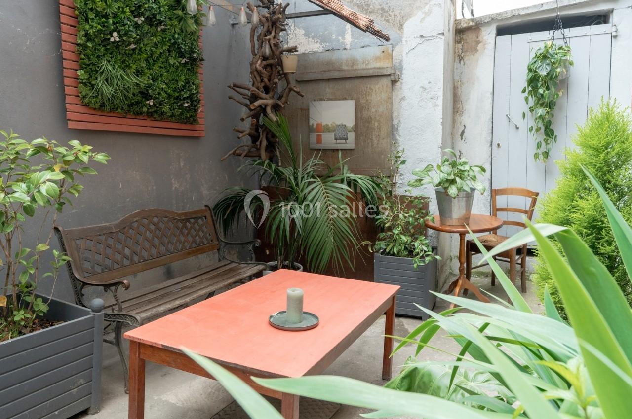 Petit patio avec banc en métal, table rouge, plantes vertes et mur végétalisé dans un cadre extérieur lumineux.