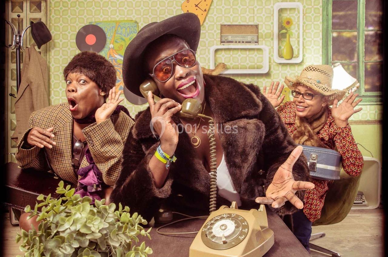 Trois personnes en tenue rétro posent de manière expressive autour d'un téléphone ancien dans une pièce au décor vintage.