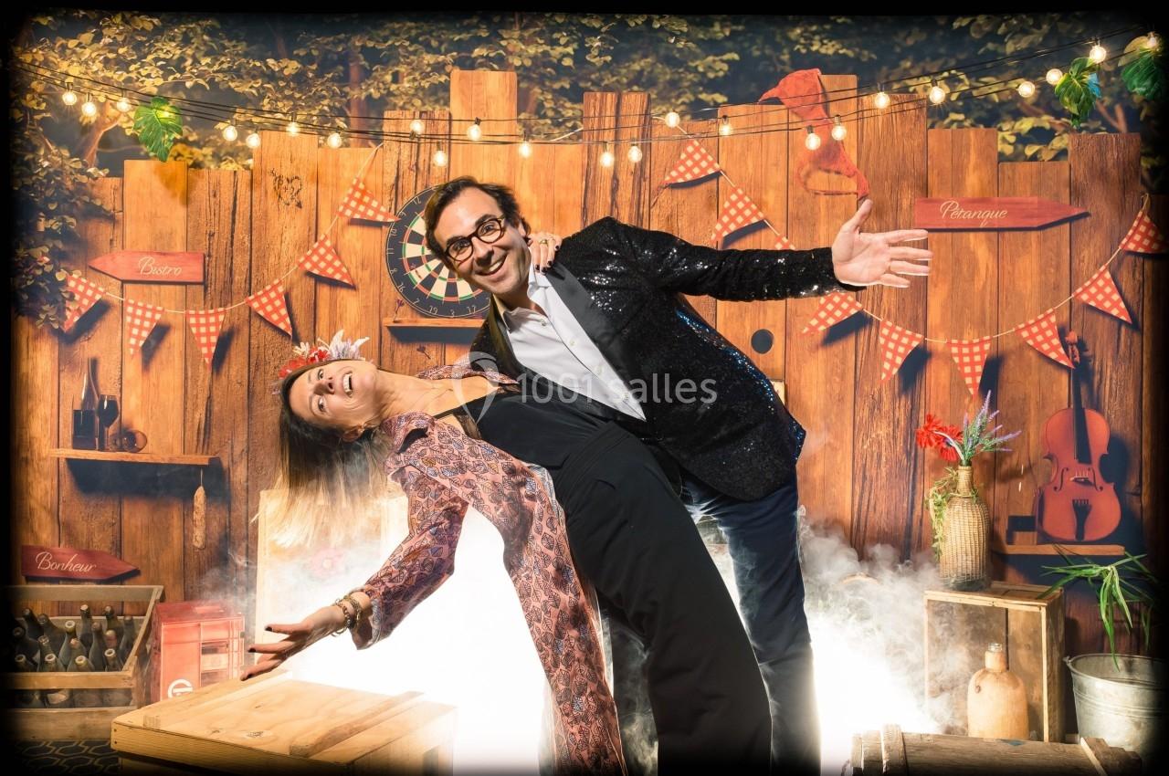 Un couple pose en dansant devant une décoration en bois avec guirlandes lumineuses et accessoires festifs.