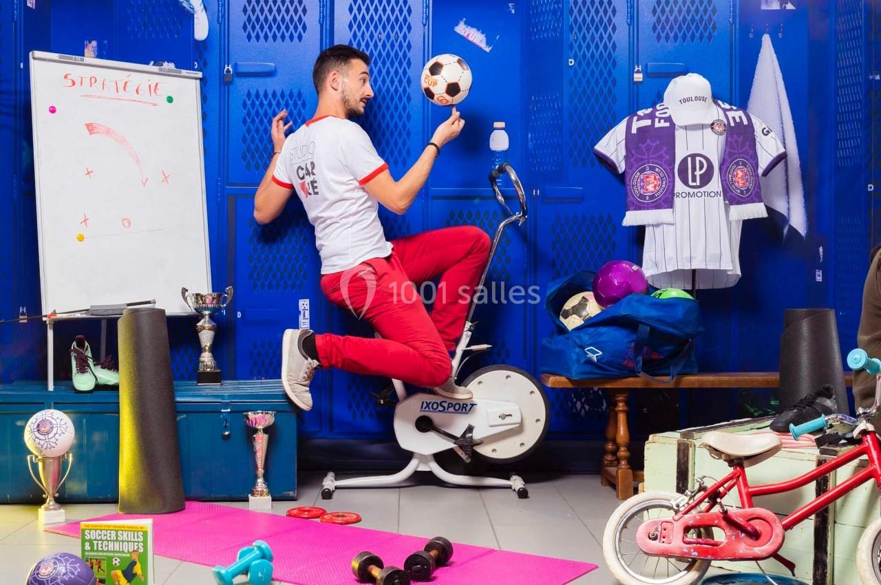 Un homme jongle avec un ballon de football sur un vélo d'appartement dans un vestiaire coloré et encombré.