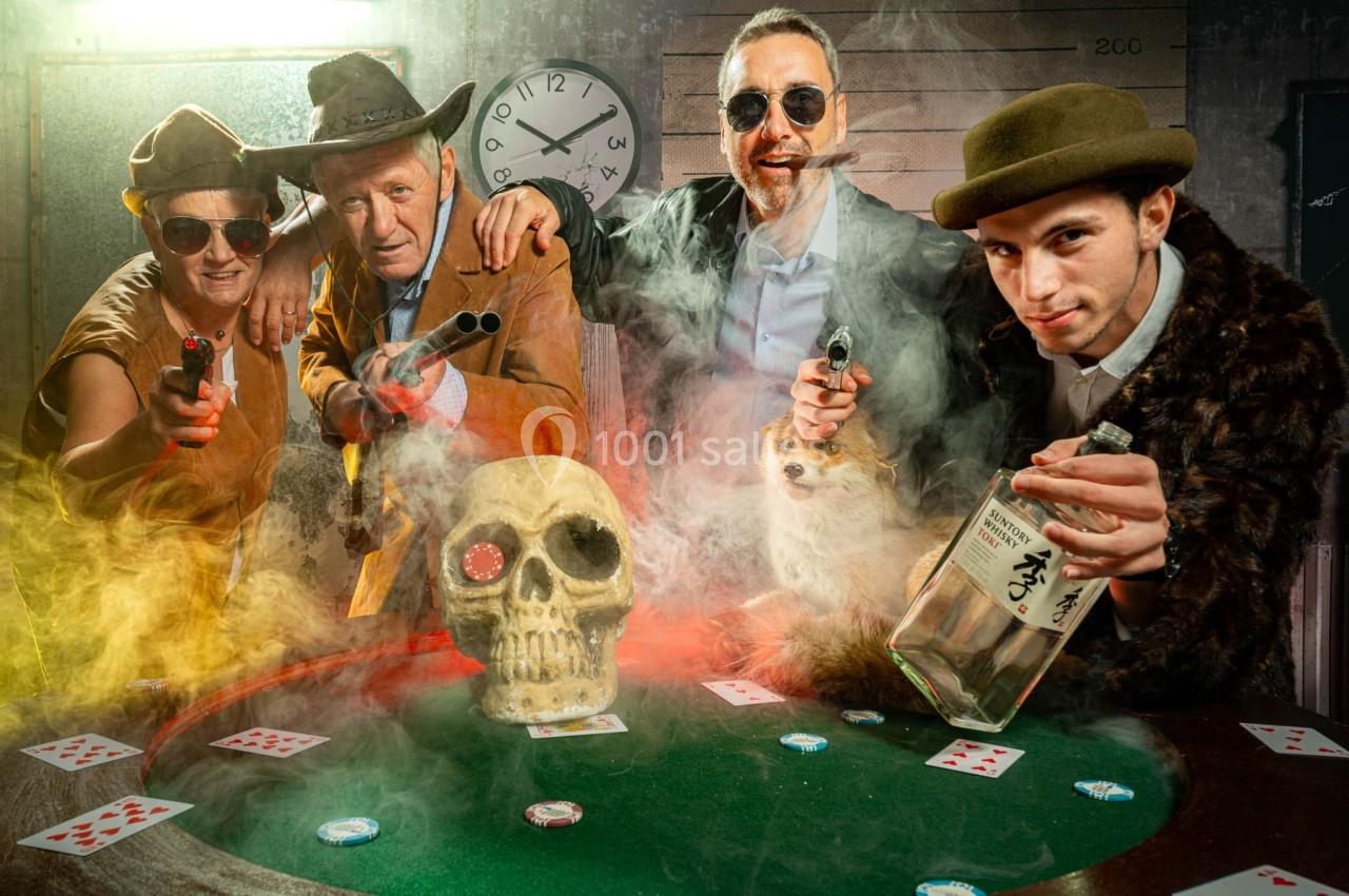 Quatre personnes déguisées autour d'une table de poker, avec un crâne, des cartes et une bouteille au premier plan.