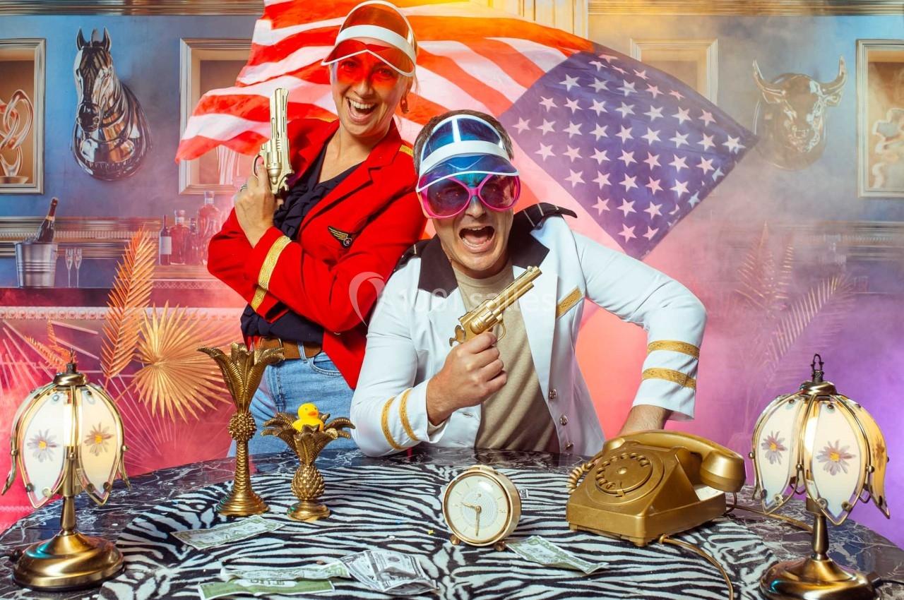 Deux personnes en tenue colorée posent avec des pistolets factices devant un décor kitsch et un drapeau américain.