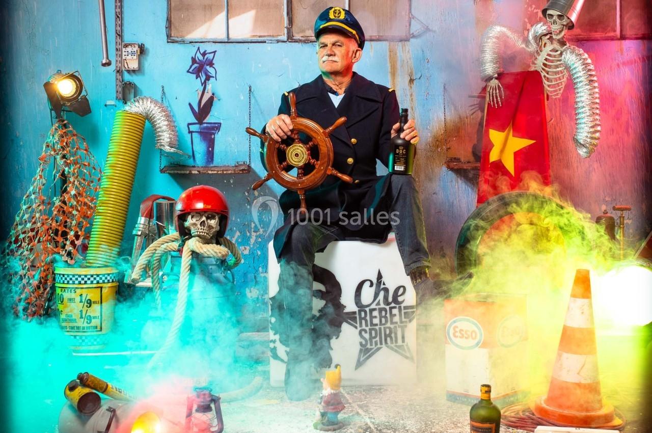 Un homme déguisé en capitaine de bateau est assis dans un décor coloré et chaotique avec des accessoires variés.