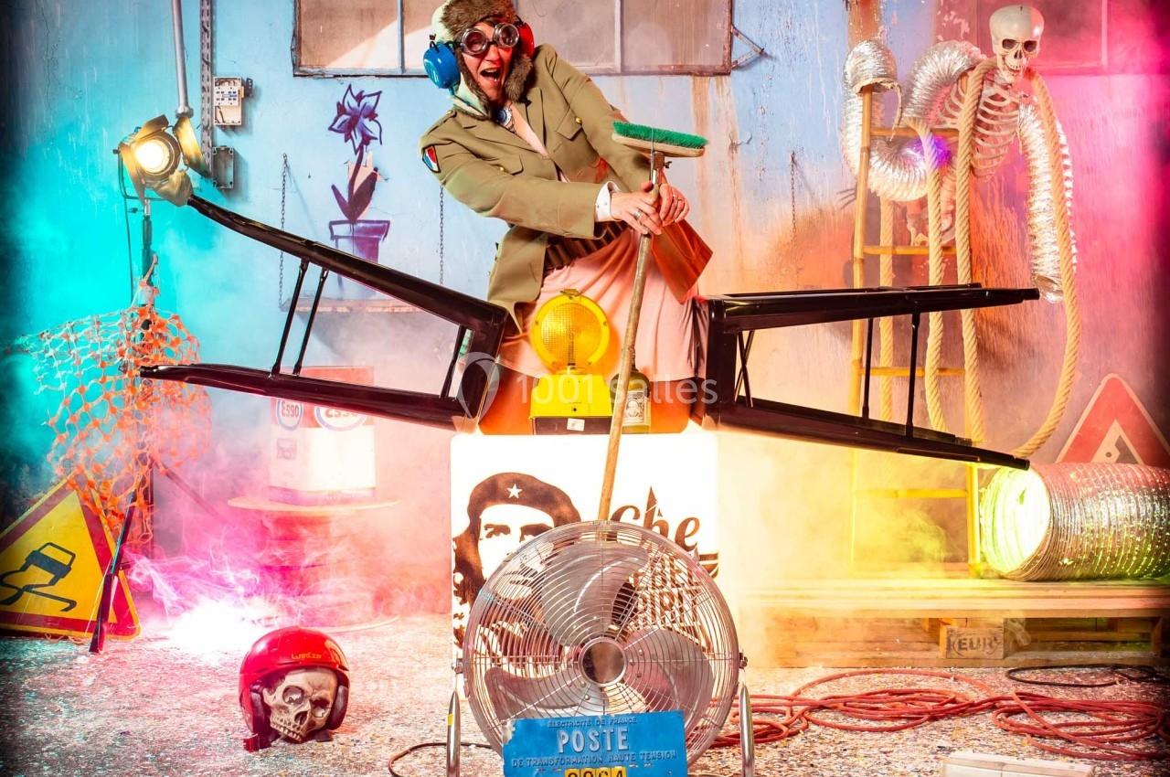 Un homme déguisé pose dans un décor coloré et chaotique, entouré d'objets variés et d'effets lumineux.