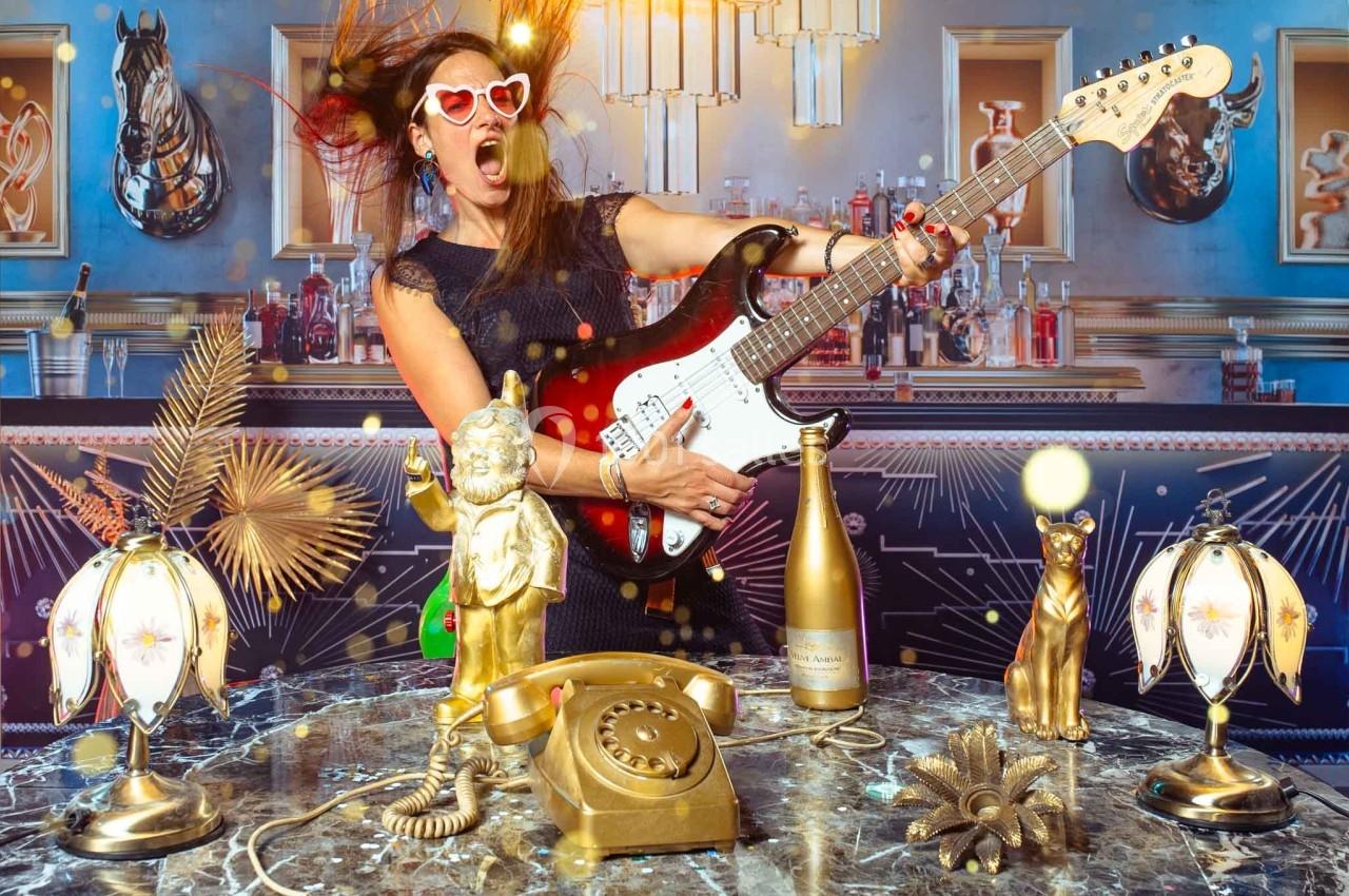 Femme portant des lunettes en forme de cœur jouant de la guitare électrique dans un décor rétro avec objets dorés.