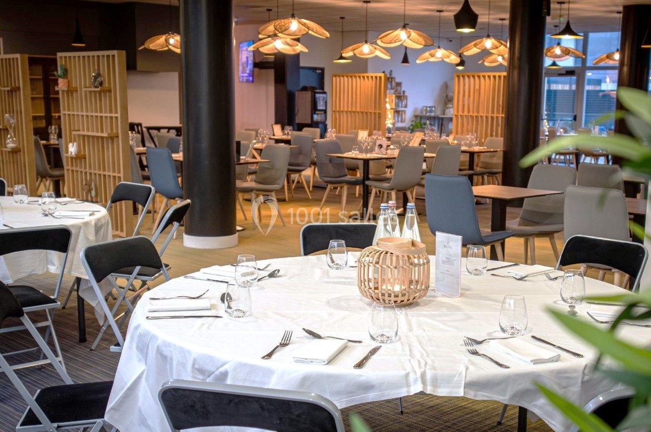 Salle de restaurant lumineuse avec tables rondes dressées, chaises noires et grises, et éclairage suspendu chaleureux.