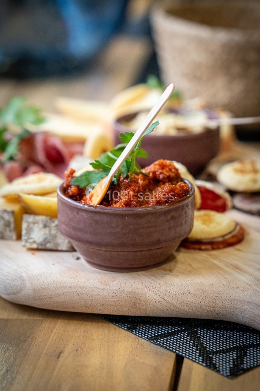 Bol de tartinade rouge avec une cuillère en bois, entouré de charcuteries, fromages et pains sur une planche en bois.