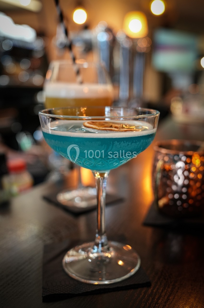 Cocktail bleu garni d'une tranche d'agrume séché, posé sur un comptoir de bar avec un autre verre en arrière-plan.