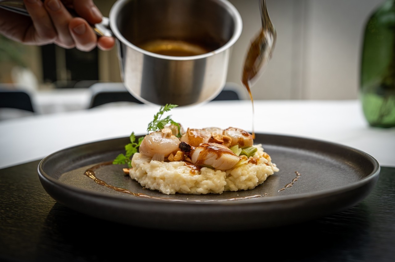 Un risotto crémeux garni de noix de Saint-Jacques, décoré de sauce versée délicatement depuis une casserole.