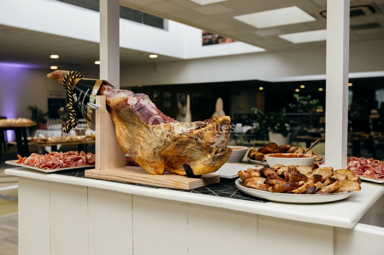 Jambon entier sur support en bois, entouré d'assiettes de charcuterie et de pain grillé dans un espace lumineux.