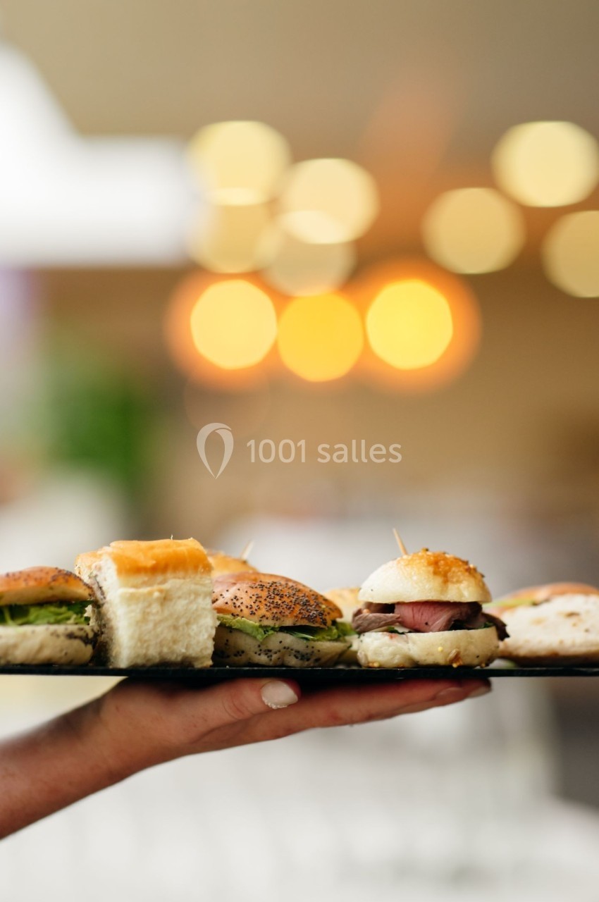 Plateau de mini-sandwichs variés tenu par une main, avec un arrière-plan flou et des lumières chaleureuses.