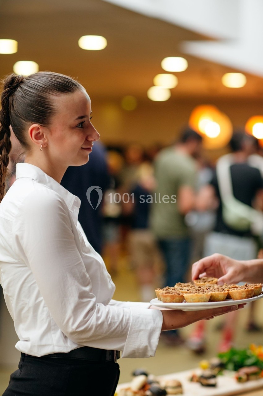 Une serveuse en chemise blanche tend un plat de desserts dans un espace intérieur animé.