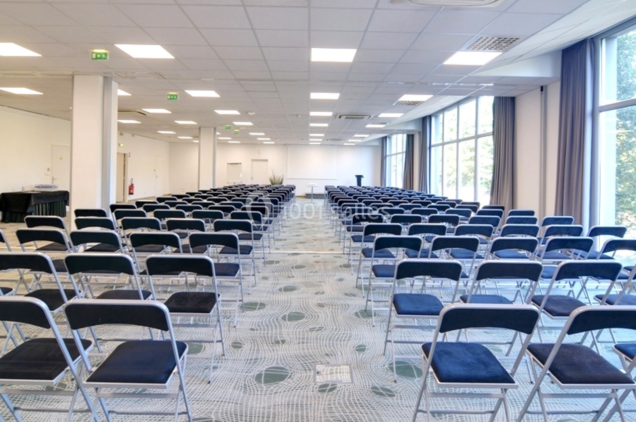 Salle de conférence lumineuse avec rangées de chaises pliantes noires alignées face à un pupitre.