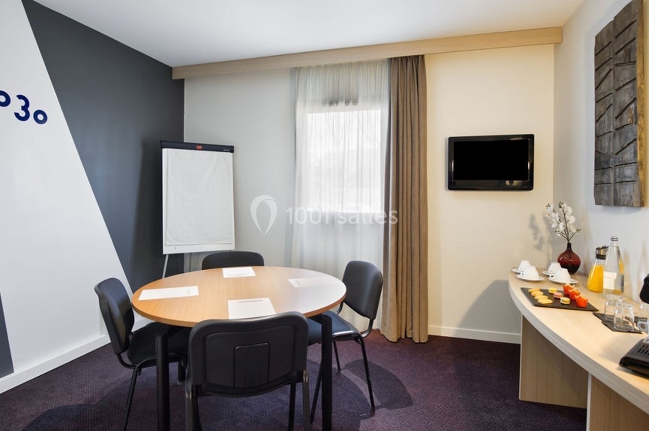 Salle de réunion avec table ronde, chaises, paperboard, écran mural, fenêtre et coin avec boissons et collations.
