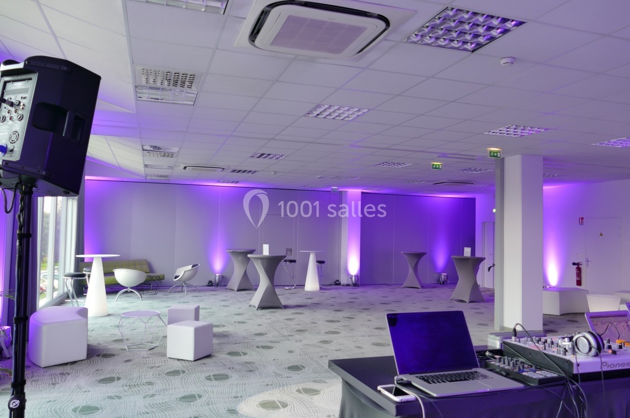 Salle de réception moderne avec éclairage violet, tables hautes, chaises design et matériel audio en premier plan.