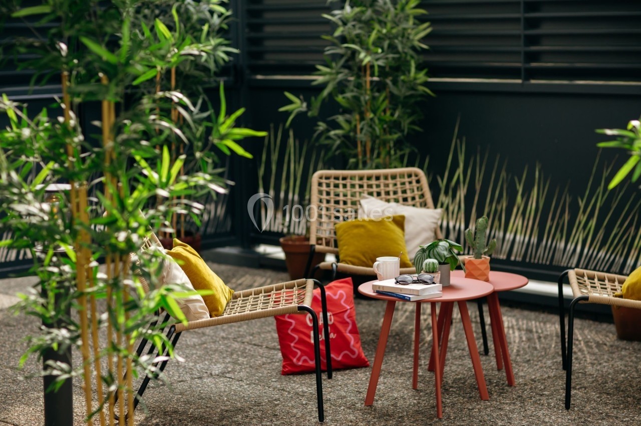 Deux fauteuils en rotin avec coussins colorés entourent une table basse rouge sur une terrasse végétalisée.