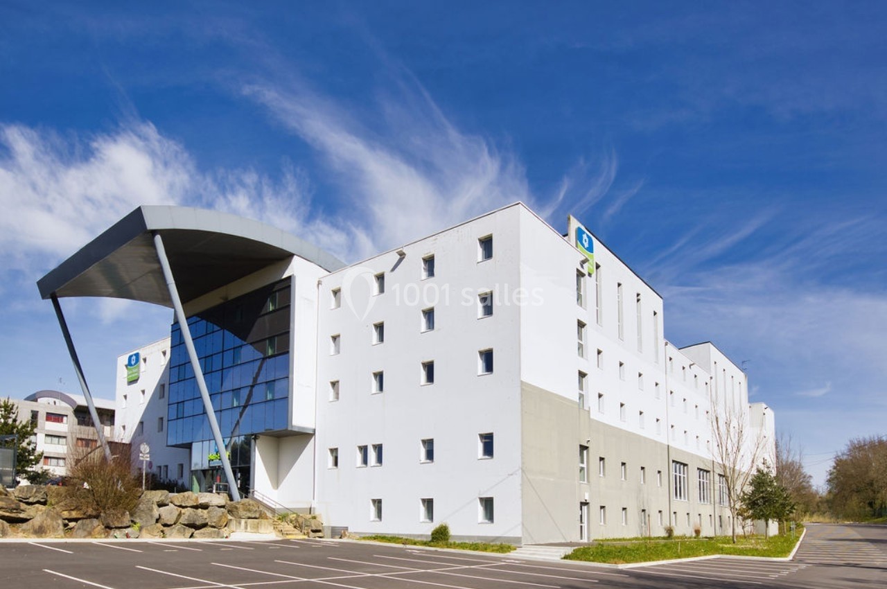 Bâtiment moderne blanc avec façade vitrée, parking vide au premier plan et ciel bleu en arrière-plan.