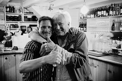 Miniature DJ Milizac (Finistère) - Florian Rion #6 Homme en chemise noire, lunettes de soleil, fumant un cigare, tenant un verre devant un bar rempli de bouteilles.