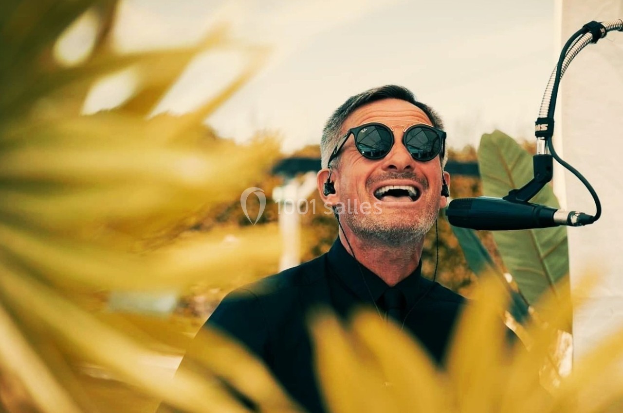 Un homme portant des lunettes de soleil chante au micro en extérieur, entouré de feuilles floues au premier plan.