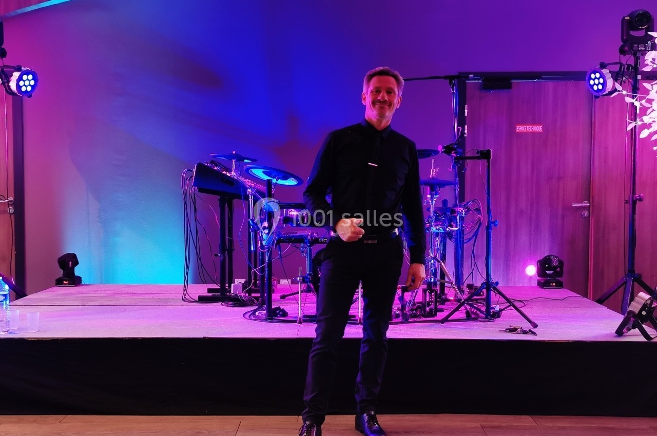 Un homme debout sur une scène éclairée en violet, entourée d'équipements de musique et de lumières décoratives.