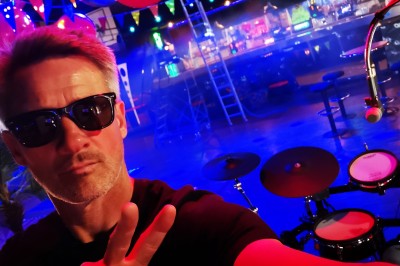 Miniature DJ Milizac (Finistère) - Florian Rion #17 Homme en chemise noire, lunettes de soleil, fumant un cigare, tenant un verre devant un bar rempli de bouteilles.