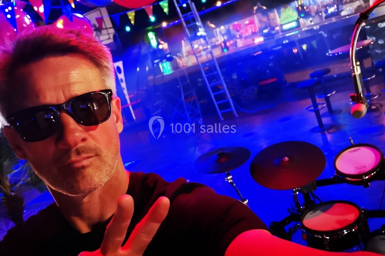 Homme portant des lunettes de soleil faisant un signe de paix devant une batterie dans une salle colorée et décorée.