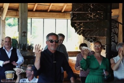 Miniature DJ Milizac (Finistère) - Florian Rion #18 Homme en chemise noire, lunettes de soleil, fumant un cigare, tenant un verre devant un bar rempli de bouteilles.