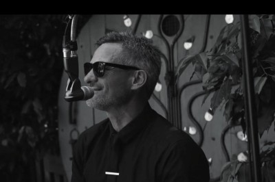 Miniature DJ Milizac (Finistère) - Florian Rion #8 Homme en chemise noire, lunettes de soleil, fumant un cigare, tenant un verre devant un bar rempli de bouteilles.