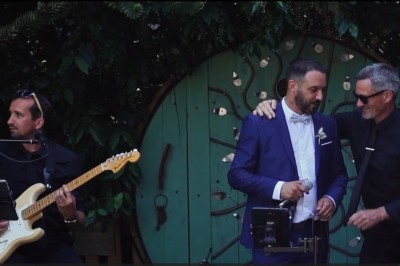 Miniature DJ Milizac (Finistère) - Florian Rion #16 Homme en chemise noire, lunettes de soleil, fumant un cigare, tenant un verre devant un bar rempli de bouteilles.