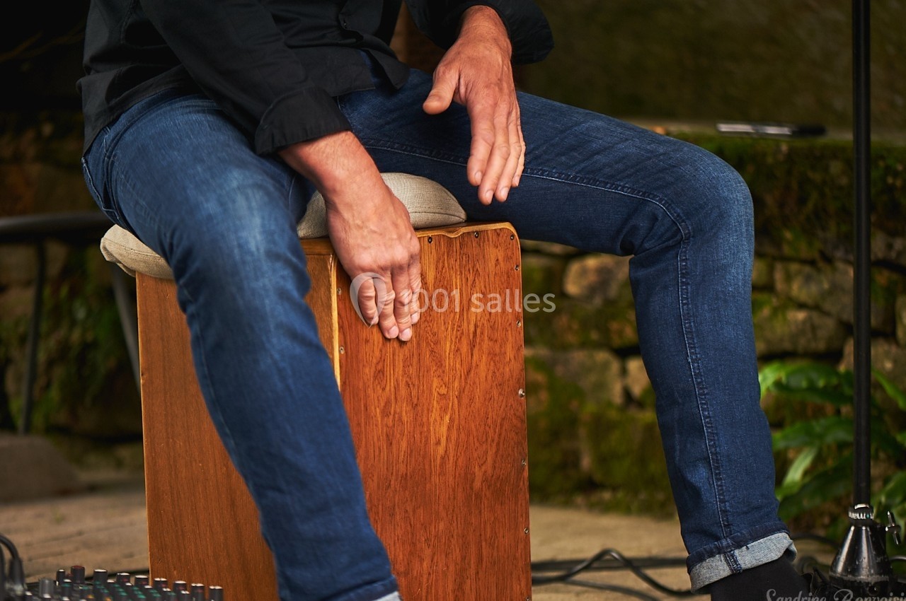 Musicien jouant du cajón en bois, ses mains frappant le devant de l'instrument dans un cadre extérieur.
