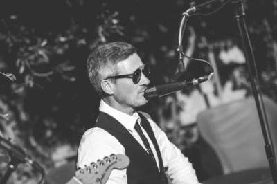 Miniature DJ Milizac (Finistère) - Florian Rion #7 Homme en chemise noire, lunettes de soleil, fumant un cigare, tenant un verre devant un bar rempli de bouteilles.