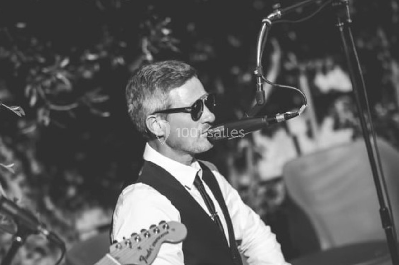 Un homme en costume et lunettes de soleil chante au micro lors d'une performance musicale en extérieur.