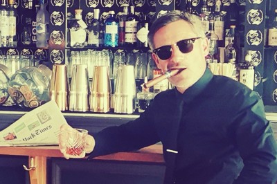 Miniature DJ Milizac (Finistère) - Florian Rion #23 Homme en chemise noire, lunettes de soleil, fumant un cigare, tenant un verre devant un bar rempli de bouteilles.