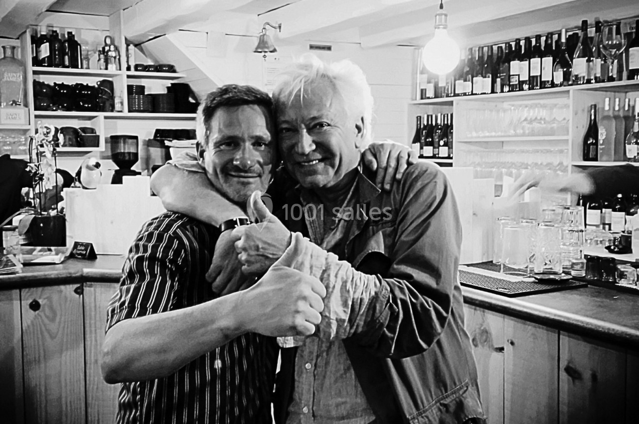 Deux hommes souriants posent ensemble dans un bar, l'un levant le pouce en signe d'approbation.