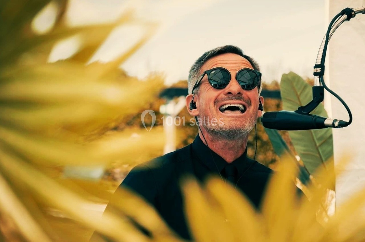 Un homme portant des lunettes de soleil chante au micro, entouré de feuilles floues au premier plan.