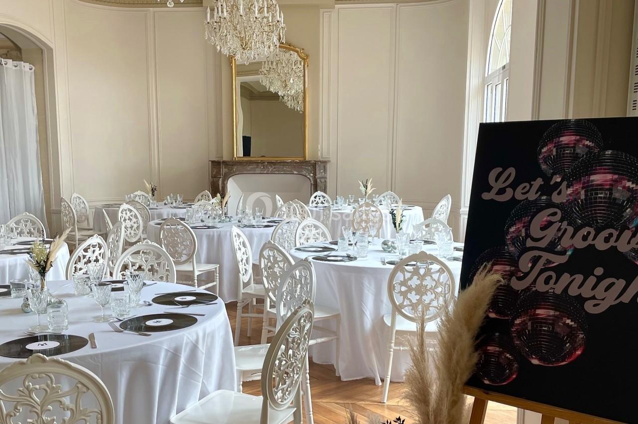 Salle de réception élégante avec tables rondes dressées, chaises blanches sculptées et panneau décoratif noir.