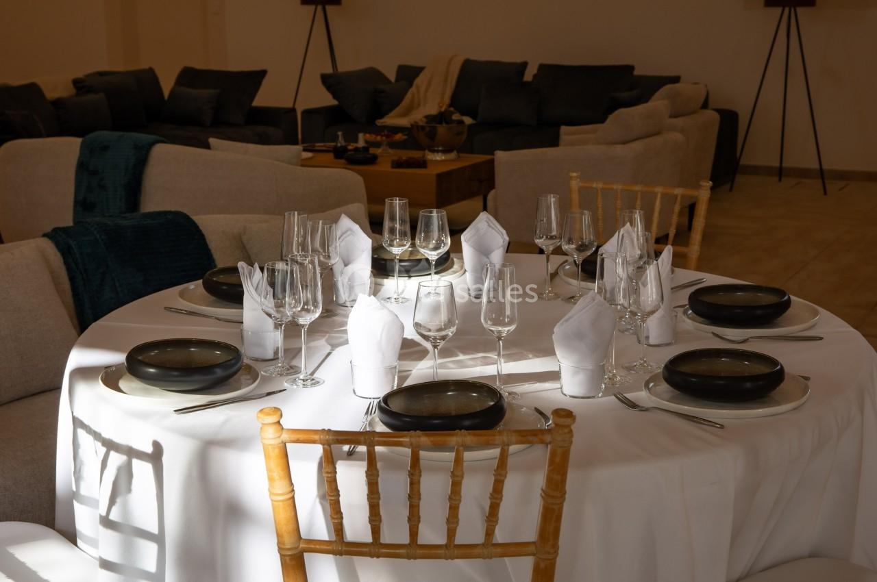 Table ronde dressée avec nappes blanches, assiettes noires, verres et serviettes pliées, entourée de chaises en bois.