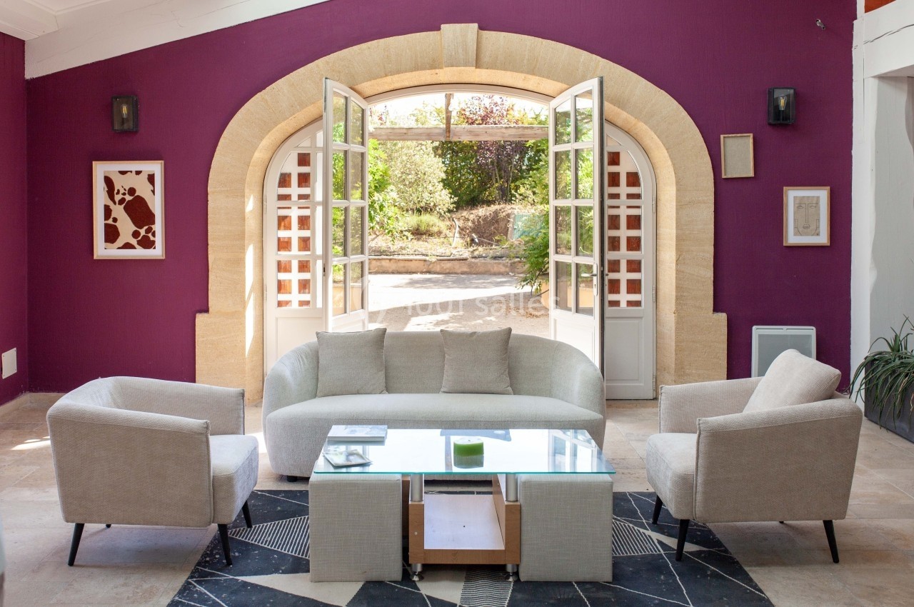 Salon lumineux avec canapé, fauteuils beiges et table basse en verre, ouvert sur un jardin par des portes-fenêtres.